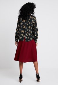 Nainen, jolla on kiharat hiukset, pukeutuu mustaan kukkakuosisessa puserossa ja polvipituisessa punaisessa hameessa, seisoen selin mustissa korkokengissä.