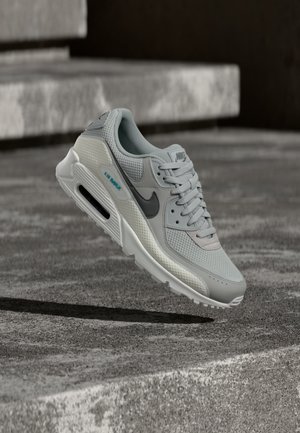 Světlé šedé tenisky Nike Air Max ve vzduchu nad betonovými schody, s texturami z meshe a kůže, černým logem a viditelnou vzduchovou podrážkou.