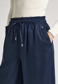 Kvinna iklädd marinblå vida byxor med elastisk midja, snörning och ficka med lock, kombinerat med en krämfärgad ribbad långärmad topp.