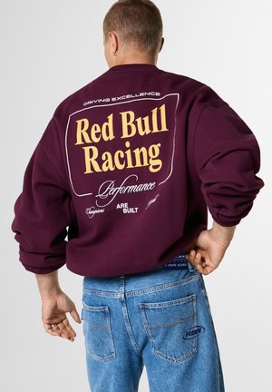 Person, die einen burgunderfarbenen Red Bull Racing Sweatshirt und blaue Jeans mit RBR-Logo trägt, von hinten vor neutralem Hintergrund gesehen.