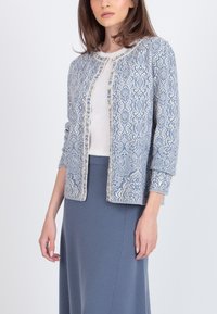 Cárdigan azul claro con patrones blancos intrincados, cuello redondo y cierre de botones. Llevado sobre una blusa blanca y una falda azul.