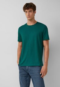 T-shirt in cotone verde con collo rotondo, maniche corte e un logo sottile sul petto. Abbinato a jeans blu.
