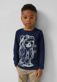 Junger Junge mit kurzen lockigen Haaren, der ein marineblaues Langarmshirt mit einem glänzenden Teddybaummotiv und beigen Hosen trägt, steht vor einer hellgrauen Wand.
