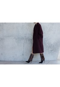 Bordowy, oversize płaszcz, teksturowany materiał; zestawiony z legginsami w panterkę i czarnymi botkami na szpilkach. Szary betonowy pejzaż w tle.