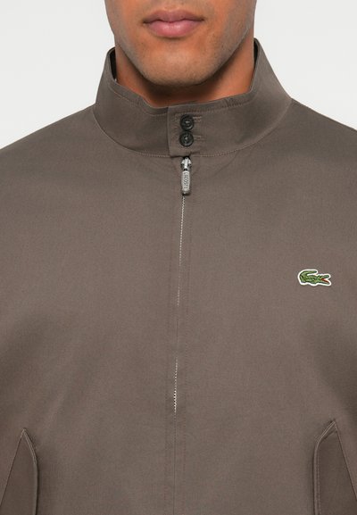 Homme portant une veste à fermeture éclair marron avec col montant, deux boutons noirs et un logo de crocodile vert sur la poitrine.