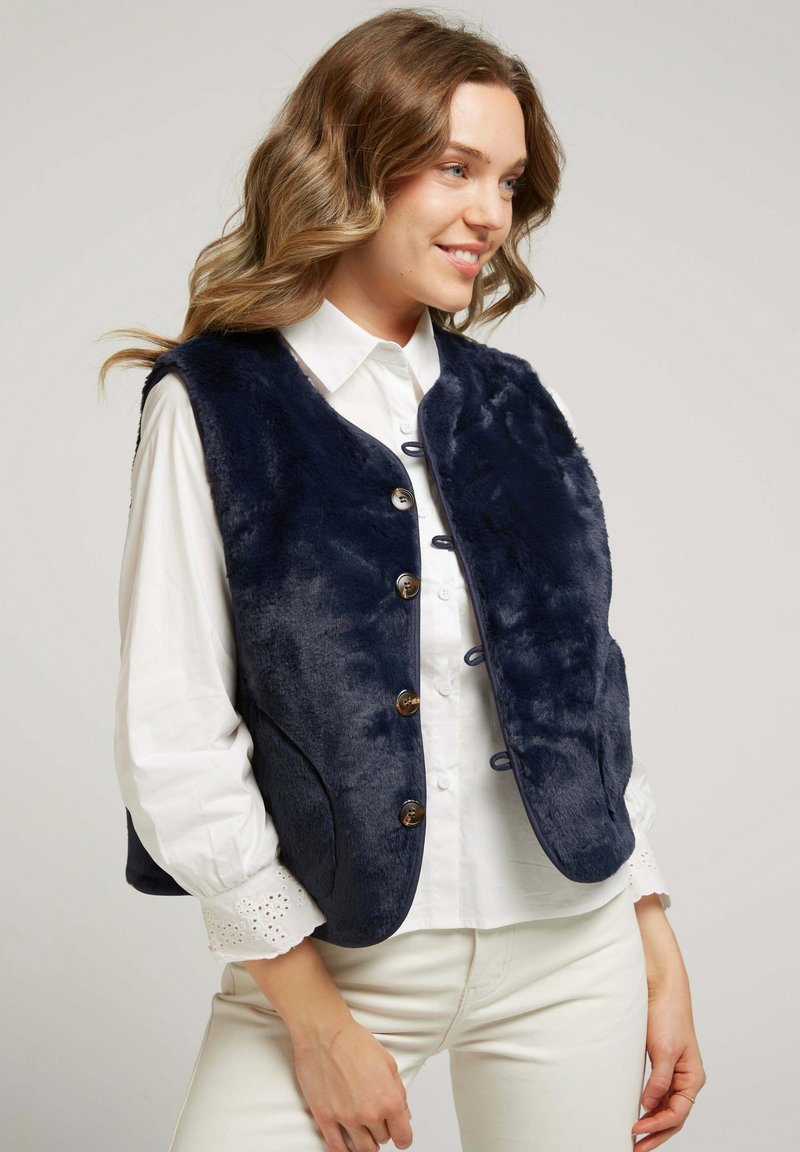 Femme portant un gilet boutonné en fausse fourrure bleu marine par-dessus une blouse blanche à manches longues avec des poignets en dentelle et un pantalon de couleur claire.