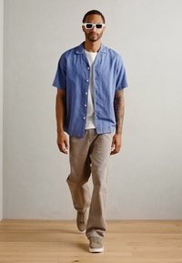 Camicia azzurra a maniche corte con bottoni, t-shirt bianca sotto, pantaloni beige chiaro, sneakers grigie e occhiali da sole bianchi. Sfondo in legno.