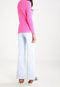 Enganliegendes rosa Ripp-Pullover, hellblaue Schlagjeans und braune Sandalen mit einer Schleifenakzent. Weiche Texturen und lebendige Farben sind prägnant.