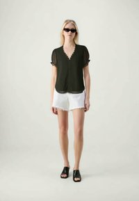 Mulher vestindo uma blusa preta com borda recortada, calções de ganga brancos desfiados, mules pretos de bico aberto e óculos de sol escuros, em fundo neutro.