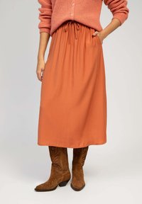 Jupe midi orange avec taille froncée et cordon de serrage. Le tissu est lisse et léger. Associée à des bottes de cowboy en daim marron avec des broderies élaborées.
