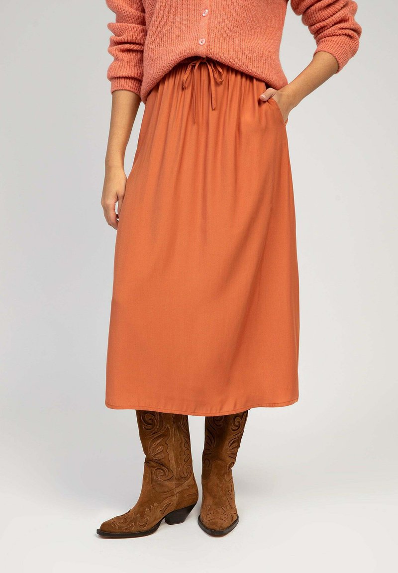 Jupe midi orange avec taille froncée et cordon de serrage. Le tissu est lisse et léger. Associée à des bottes de cowboy en daim marron avec des broderies élaborées.