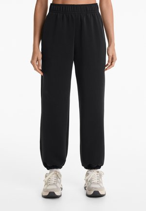 Zwarte trainingsbroek met een elastische tailleband, taps toelopende boorden en zijzakken, gecombineerd met grijze en witte sneakers. Glad stoffen textuur.