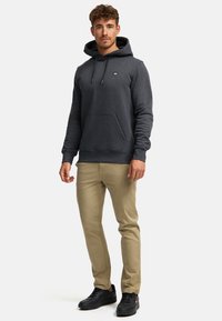 Homme debout portant un sweat à capuche gris foncé, un pantalon beige et des baskets noires sur un fond blanc uni.