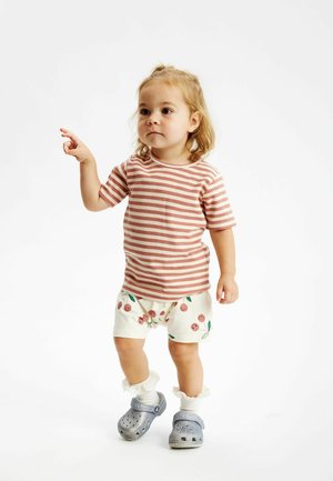 Chemise à manches courtes rayée en rose et blanc, associée à un short crème avec des motifs de cerise. Crocs gris avec des chaussettes blanches à volants.