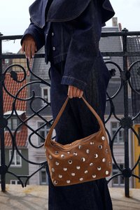 Borsa a mano in suede marrone con accenti circolari in tonalità argento, texture morbida, chiusura a zip e forma curva, portata da una persona in abbigliamento di denim.