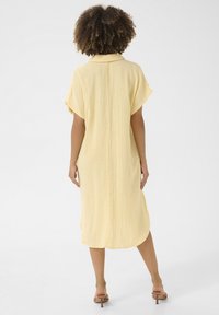 Abito camicia giallo corto con maniche corte, realizzato in tessuto leggero, con colletto e spacchi laterali. Vista posteriore mostrata.