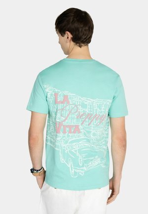 Homme en t-shirt turquoise avec dessin linéaire blanc d'une voiture vintage et d'un paysage urbain, texte rose "LA DOLCE VITA" au dos, portant un short blanc.