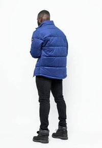 Blå, vadderad jacka med hög krage, tillverkad av mjukt material, med horisontell sömnad och en liten logotyp på vänster axel.