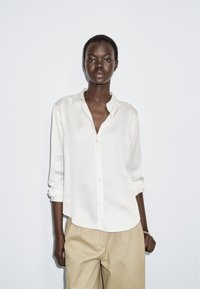 White button-up shirt with a collar and long sleeves, paired with beige trousers. Smooth fabric, relaxed fit, and subtle sheen.