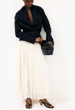 Kobieta w czarnej bluzce z głębokim dekoltem w serek, długiej plisowanej kremowej spódnicy, czarnych sandałach na obcasie, trzymająca czarną skórzaną torebkę.