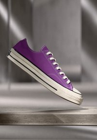 Lilla lerretssko med hvit gummisåle, rund tå og hvite snørebånd. Har en svart stripe langs sålen og teksturert tåhette.