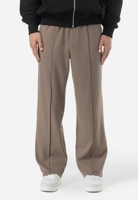 Pantalons beige à-large avec une finition lisse et texturée. Présente des coutures centrales et portés avec des baskets blanches. Veste zippée noire en dessus.