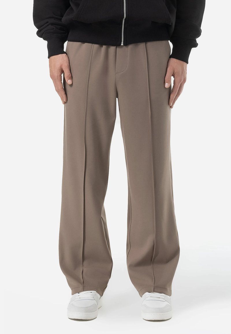 Pantalons beige à-large avec une finition lisse et texturée. Présente des coutures centrales et portés avec des baskets blanches. Veste zippée noire en dessus.