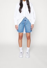 Witte blouse met lange mouwen en knopen, blauwe denim shorts met rafelige zoom, witte enkelsokken en grijze sneakers met zwarte accenten.