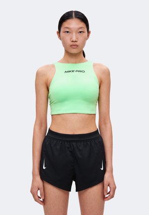 Jeune femme portant un crop top Nike Pro vert clair et un short de course Nike noir, debout devant un fond uni.