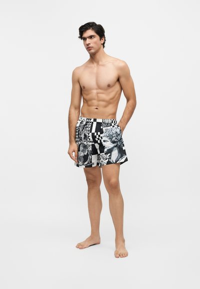 Homme torse nu, debout pieds nus, portant un short à motifs noirs et blancs avec des sculptures classiques et des motifs en damier sur un fond uni.
