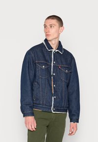 Jeansowa kurtka w ciemnoniebieskim kolorze z białym wykończeniem z polaru, oferująca klasyczny design, zapięcie na guziki oraz dwie kieszenie na piersi z kontrastowym przeszyciem.