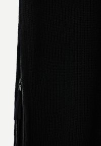 Svart stickad plagg med ett strukturerat mönster och en siddragkedjedetalj. Tyget är tjockt och tungt, vilket ger värme.