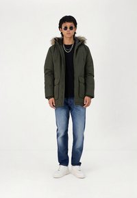 Oljčno zelena podložena jakna z notranjo obrobo iz krzna, z dvema sprednjima žepoma, nošena čez črno majico, kombinirana z modrimi kavbojkami in belimi supergami.