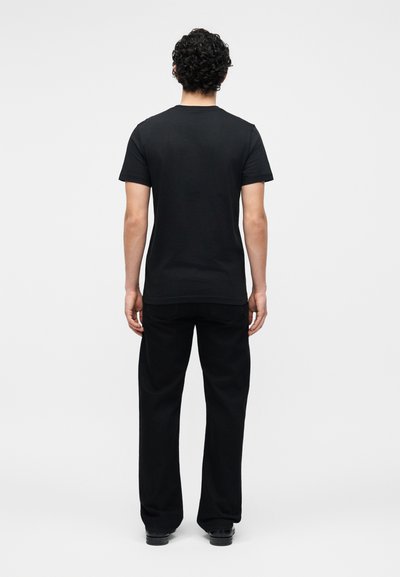 Homme aux cheveux noirs et bouclés, debout de dos, portant un t-shirt noir uni à manches courtes, un pantalon noir et des chaussures noires sur fond blanc.
