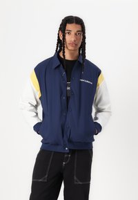 Veste bleu marine avec des manches jaunes et blanches, boutons-pression et logo "new balance". Elle a une coupe décontractée et deux poches latérales.