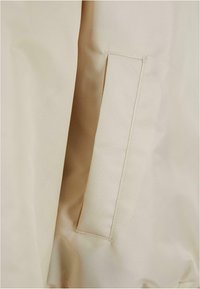 Beige Stoff mit glatter Textur, der eine seitliche Tasche aufweist, die von einfacher Naht umrandet ist. Die Tasche ist rechteckig und unauffällig.