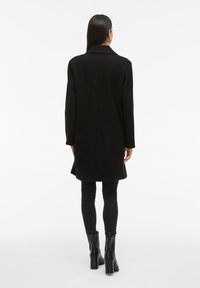 Cappotto di lana nero con una silhouette lunga, colletto a revers e taglio dritto. Il modello indossa pantaloni neri aderenti e stivaletti con tacco.