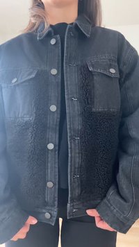 Person trägt eine schwarze Jeansjacke mit Shearling-Einsätzen, silbernen Druckknöpfen und Klappentaschen auf der Brust, steht drinnen.