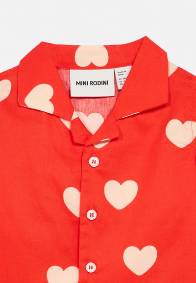 Chemise rouge à manches courtes avec de grands motifs en forme de cœur crème. Comprend un col et cinq boutons blancs, étiquetée "MINI RODINI."
