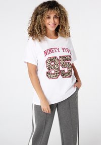 Femme souriante avec des cheveux bouclés portant un t-shirt blanc avec "Ninety Five 95" en imprimé léopard et un pantalon de survêtement gris avec des rayures blanches sur les côtés.