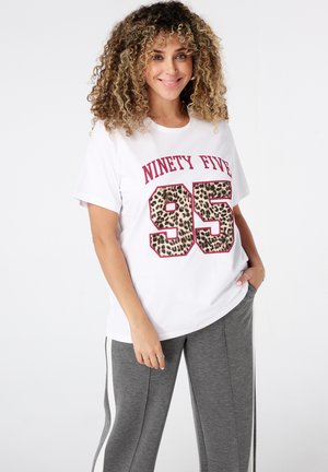 Glückliche Frau mit lockigem Haar, die ein weißes T-Shirt mit "Ninety Five 95" in Leopardenmuster und graue Sweatpants mit weißen Seitestreifen trägt.