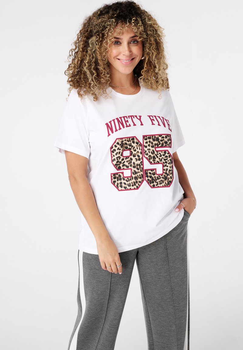 Femme souriante avec des cheveux bouclés portant un t-shirt blanc avec "Ninety Five 95" en imprimé léopard et un pantalon de survêtement gris avec des rayures blanches sur les côtés.