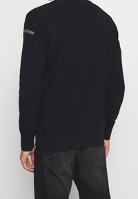 Pull en tricot bleu marine avec un motif texturé, poignets et ourlet côtelés, orné d'un patch logo sur l'épaule.