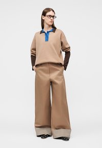 Donna che indossa pantaloni beige a gamba larga con risvolti a quadri, top a maniche lunghe beige e blu, scarpe nere e occhiali, in piedi con le mani nelle tasche.