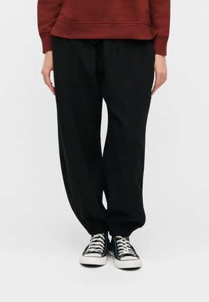 Άτομο που φοράει μαύρο φαρδύ παντελόνι με ελαστικές μανσέτες, μαύρα και λευκά αθλητικά παπούτσια και μακρυμάνικο τοπ σε σκουριά απόχρωση, στέκεται σε απλό φόντο.