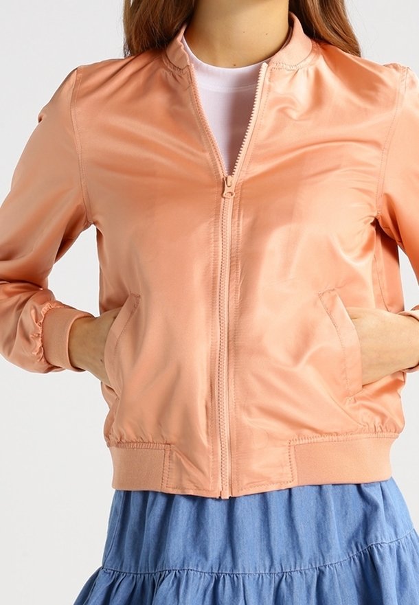 Femme portant une veste bomber en satin pêche clair avec poches avant, fermeture éclair et poignets élastiques, par-dessus un haut blanc et une jupe bleue.