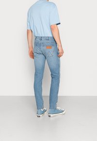 Modré džíny s úzkým střihem, s hnědou koženou nášivkou na zadním pásu a bílými bočními pruhy, v kombinaci se světle modrým tričkem a modrými teniskami.