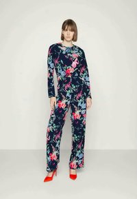 Macacão floral em tecido azul-marinho com padrões vibrantes em rosa e verde, mangas longas, corte descontraído e sapatos de salto alto vermelhos. Textura suave e decote redondo.