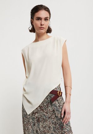 Vrouw met kort bruin haar die een mouwloos wit topje en een rok met motief draagt, met een bordeauxrode riem en gouden gesp.