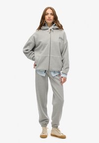 Grauer Kapuzen-Zip-Hoodie mit Kordelzügen und Fronttaschen, kombiniert mit passenden grauen Jogginghosen, getragen über einem blauen Shirt. Beigefarbene Sneakers.